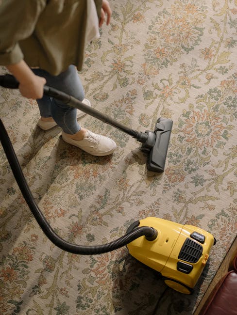 A person operating a yellow portable carpet cleaner with a black handle and vent, positioned on a patterned carpet in a room with beige and greenish tones. The scene shows the appliance's control pane